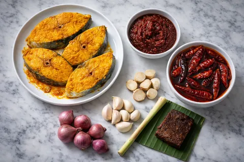 Ikan tenggiri disalin dengan kunyit hidup tumbuk dan garam dalam dulang, bersama rempah paste merah tua yang baru dikisar dalam blender — cili kering, bawang, belacan
