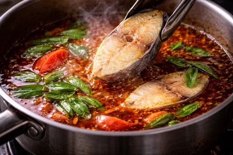 Ketul ikan tenggiri disusun perlahan ke dalam kuah asam pedas merah yang mendidih, daun kesum segar hijau masuk akhir, tomato merah dan bendi sudah masak