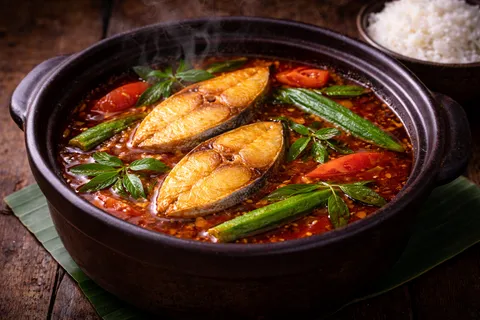 Asam Pedas Ikan Malaysia siap dalam periuk tanah — kuah merah-oren pekat berkilat dengan minyak merah terapung, ikan tenggiri utuh, bendi hijau, tomato, dan daun kesum segar mengapung