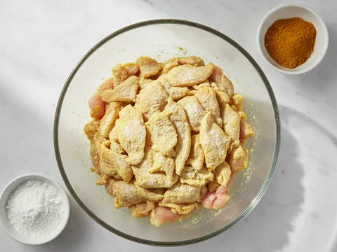 Hirisan isi ayam nipis digaul dengan tepung jagung dan serbuk kunyit dalam mangkuk