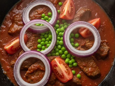Bawang besar cincin, tomato belah empat dan kacang pis dimasukkan ke atas daging masak merah dalam kuali