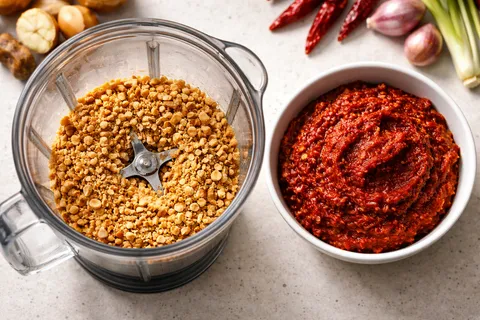 Kacang tanah sangai dikisar kasar dalam blender — tekstur serpihan kasar, bukan halus seperti butter; pes rempah merah-oren dari cili kering dan rempah dalam mangkuk berasingan