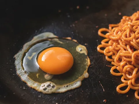 Telur dipecahkan di tepi wok bersebelahan mee goreng merah, kuning telur masih pecah separa masak