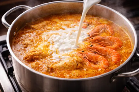 Santan putih pekat dituang perlahan ke dalam kuah kari yang sedang mendidih, udang merah-oren segar baru dimasukkan, kuah berubah jadi kuning-oren berkrim