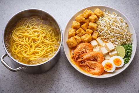 Mee kuning segar dicelur dalam periuk air mendidih, taugeh putih segar di tepi, telur rebus dibelah dua menunjukkan kuning telur yang masak sempurna, tauhu goreng keemasan dihiris